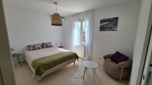 Et soveværelse med en seng, en stol, et bord, en hængende lampe, et vindue med gardiner og et billede på væggen.