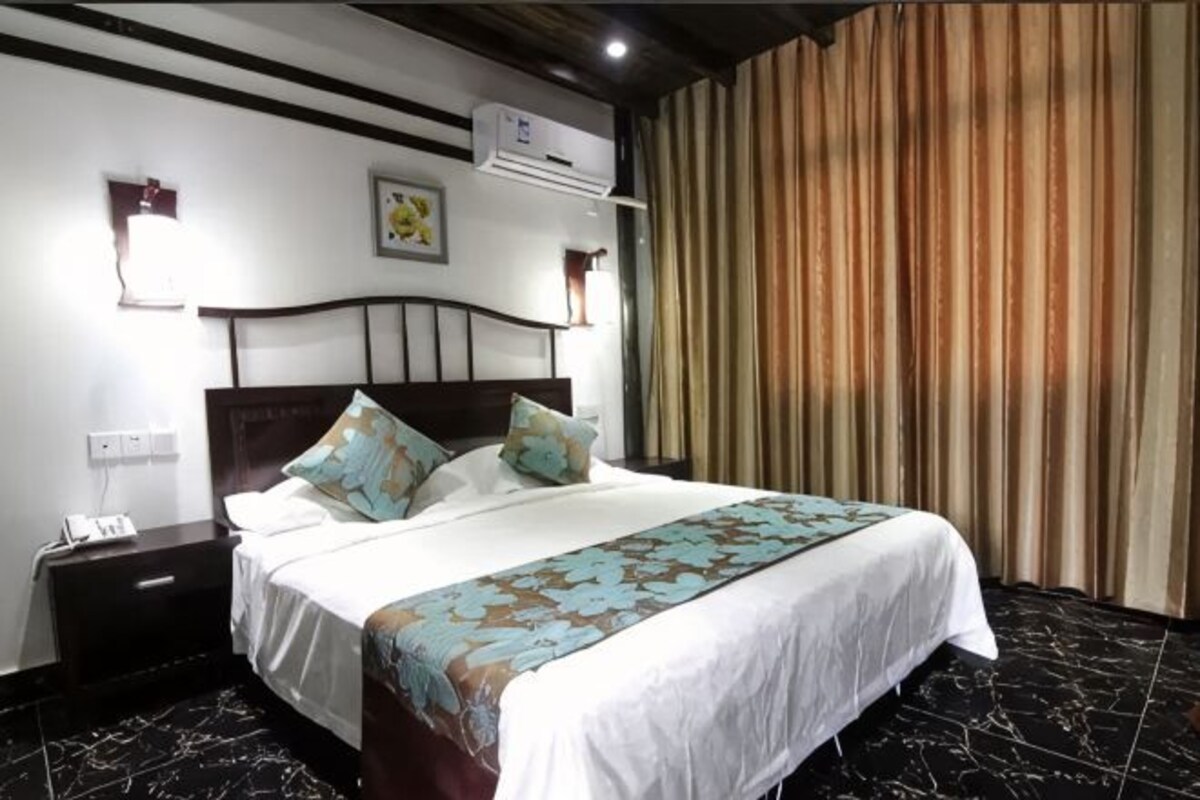 A hotel room with a bed, nightstand, wall-mounted air conditioning unit, and a curtain.