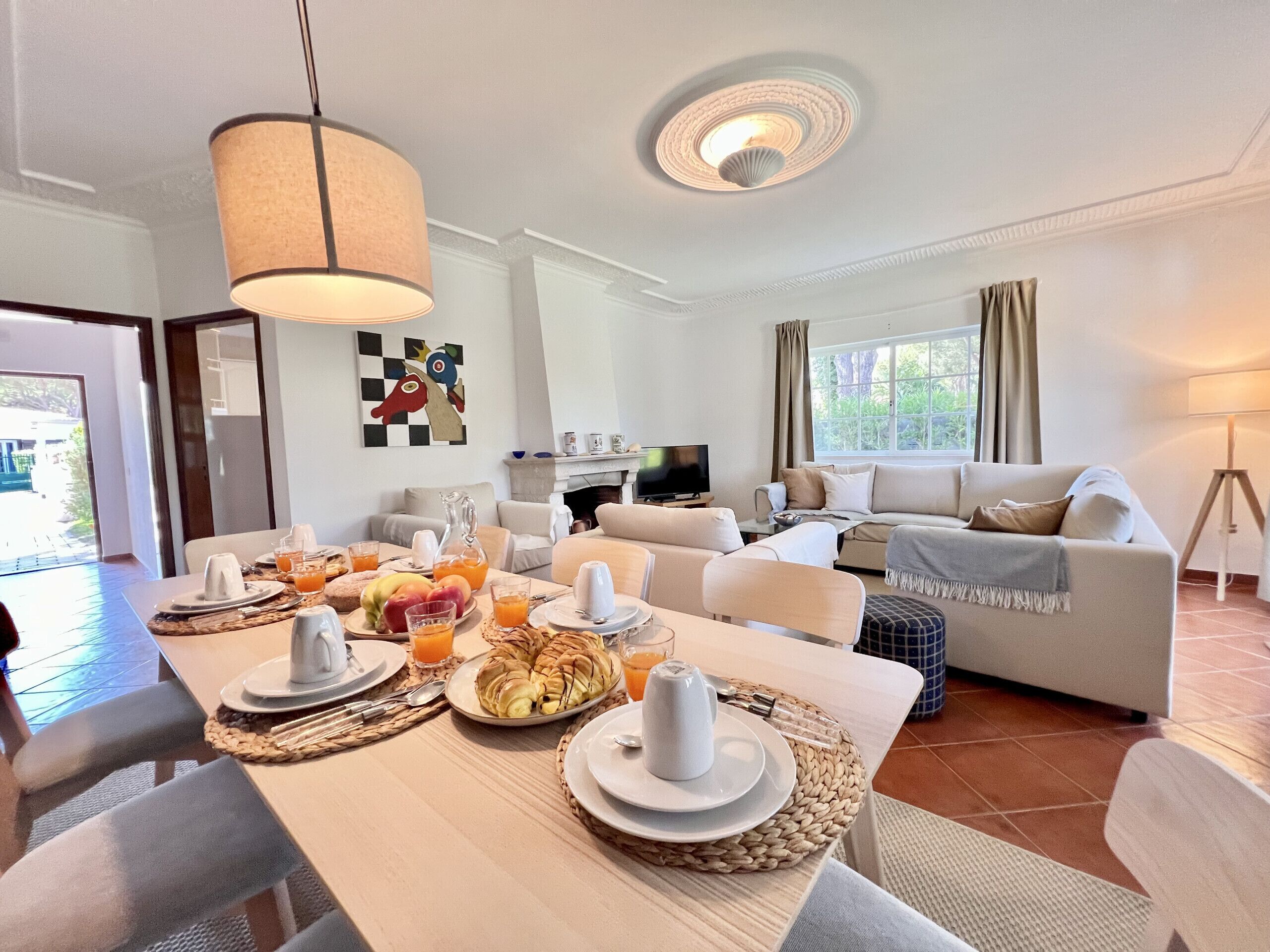 A well-lit living room with a dining area set for breakfast, featuring a sofa, television, and a view of the outdoors.