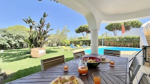 Eine Terrasse mit Frühstückstisch, inklusive Croissants und Saft, mit Blick auf ein Schwimmbad und einen gepflegten Garten.