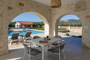 An outdoor patio with a pool, stone arches, and a view of a house and mountains. - Villa Chara with Heated Pool, by Estia (Achlades)