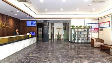 Zona de recepción de hotel con suelo de mármol, mostrador de recepción, pantalla de televisión y área de estar con un sofá y una pequeña mesa.
