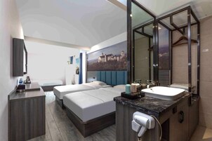 Una habitación de hotel moderna con una cama grande, una zona de estar separada, un baño con un espejo amplio y un cuadro de paisaje urbano en la pared.