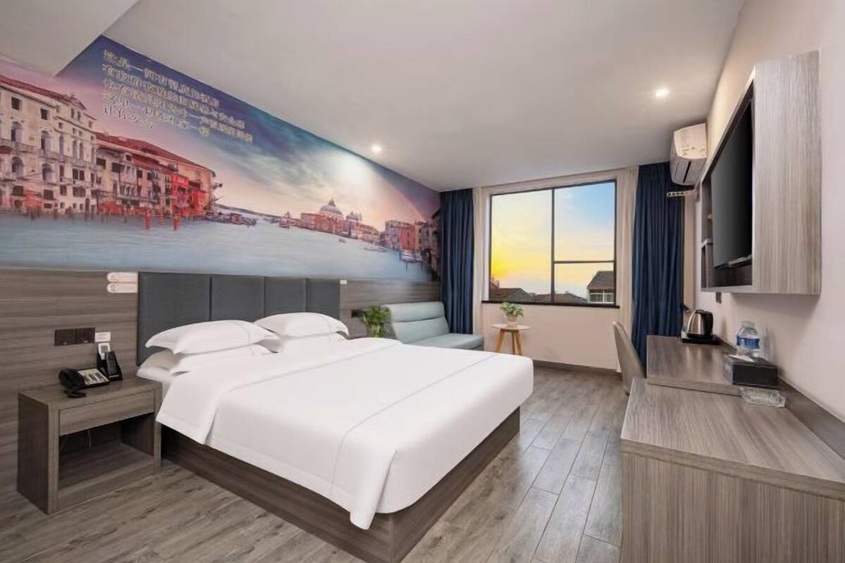 Ein Hotelzimmer mit einem großen Bett, einem Schreibtisch, einem Sessel, einem kleinen Tisch, einem Fernseher und einem Fenster mit Vorhängen.