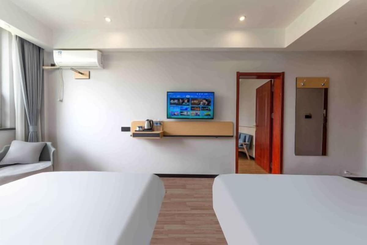 A hotel room with two beds, a TV mounted on the wall, a small shelf with a coffee maker, and a wooden floor.