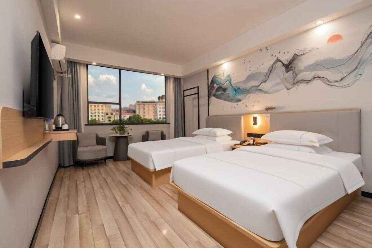 Ein Hotelzimmer mit zwei Betten, einem Fernseher, einem Schreibtisch mit Stuhl, einem Fenster mit Stadtblick und einem Bild an der Wand.