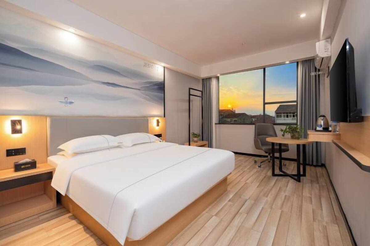 Ein Hotelzimmer mit Bett, Nachttisch, Schreibtisch, Stuhl, Fernseher und einem großen Fenster mit Sonnenuntergangsblick.