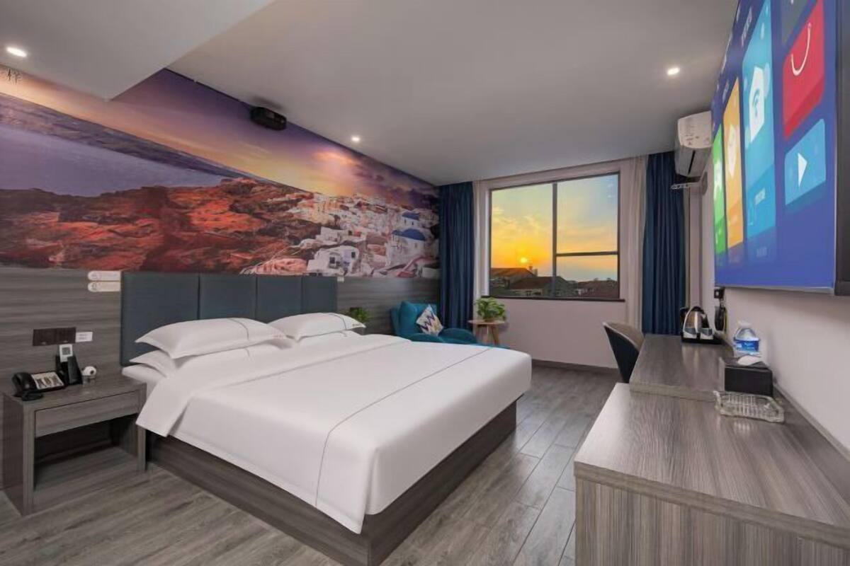 Ein Hotelzimmer mit Bett, Schreibtisch, Stuhl, Nachttisch, Fenster mit Sonnenuntergangsblick und Wandgemälde.