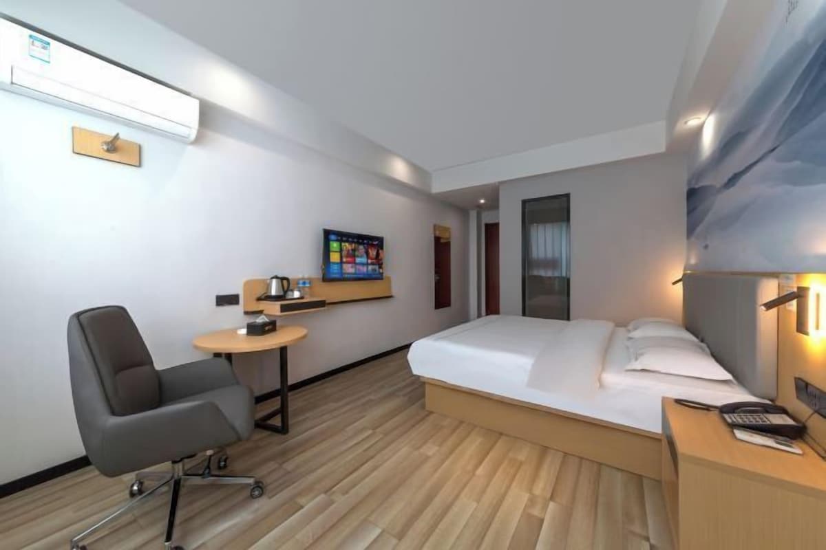 A modern hotel room with a bed, a desk, a chair, a small table, and a wall-mounted TV.