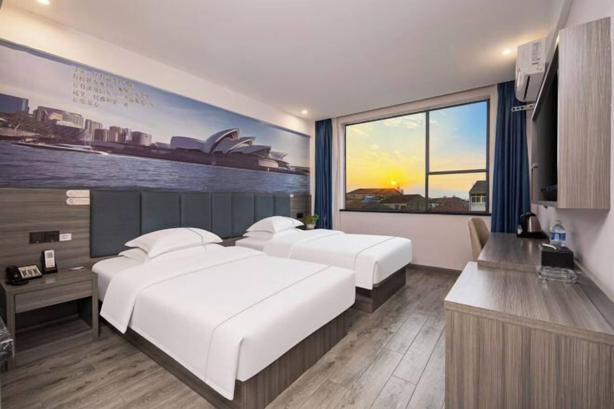 Ein Hotelzimmer mit zwei Betten, einem Schreibtisch, einem großen Fenster mit Sonnenuntergangsblick und einem Stadtbild-Wandgemälde.