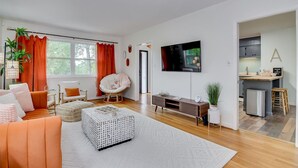 Un salon moderne comprenant un canapé, un fauteuil et un pouf, un téléviseur à écran plat et un coin cuisine avec un panneau indicateur.