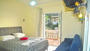 A bedroom with a bed, a sofa, a table, and a balcony with a view of buildings and greenery. - Holiday House Letta Very Quiet Location Agios Gordios Beach (Corfu)
