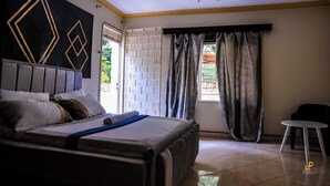 A bedroom with a bed, a window with curtains, a small table, and a wall with geometric patterns. - Golden Court (Kampala)