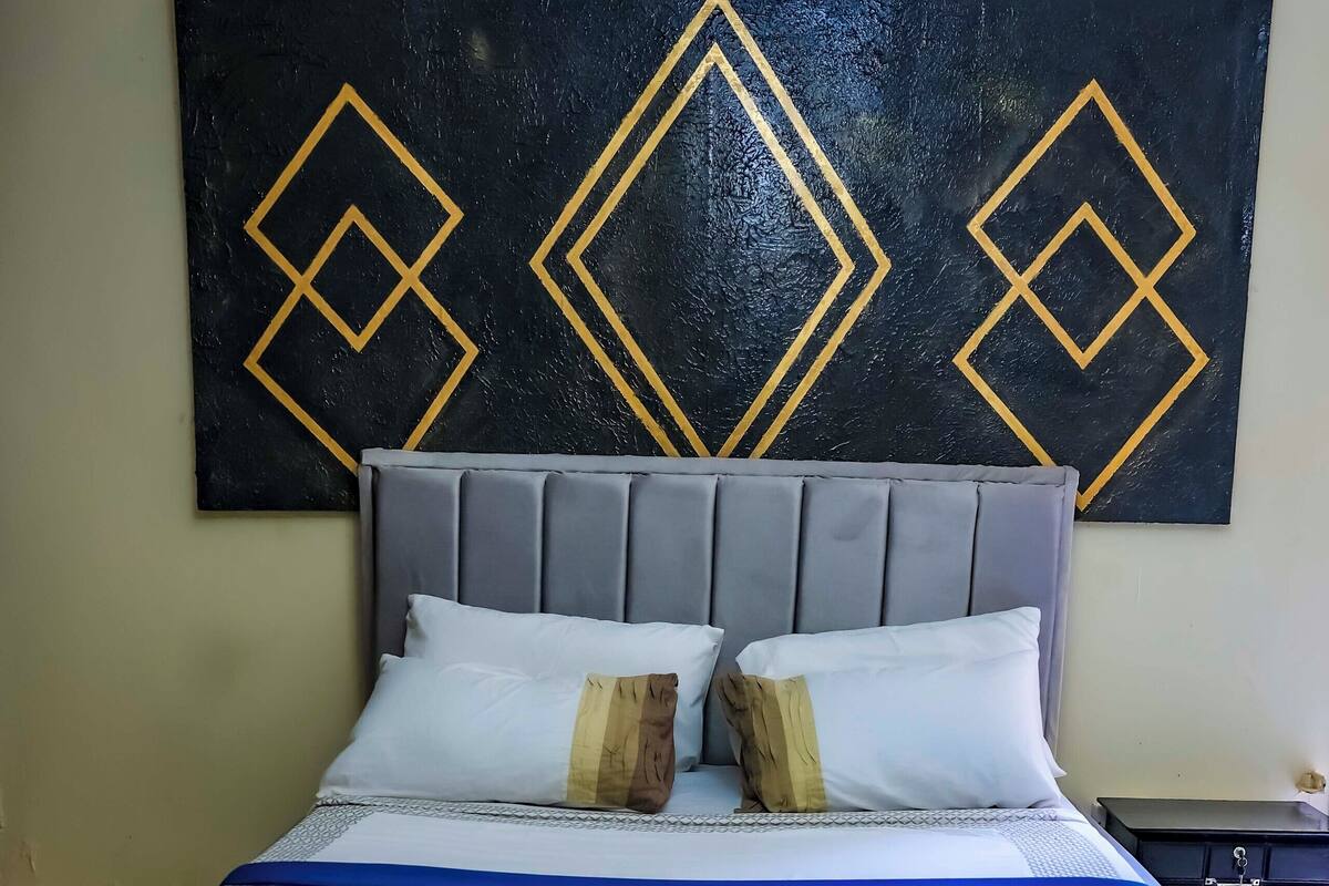 Cama bem arrumada com cabeceira cinza, roupa de cama branca e dois blocos de madeira como almofadas decorativas.