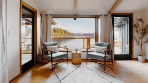 A modern living room with two grey armchairs, a wooden coffee table, and a large window offering a view of a lake and forest.