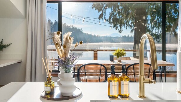 A modern kitchen with a large window overlooking a lake, a dining area with outdoor seating, and a view of mountains.