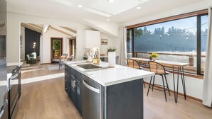 A modern kitchen with a large island, wooden flooring, and a dining area with a view of trees through large windows.