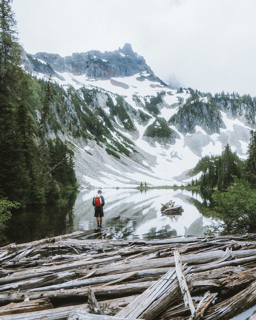 A person standing on a log over a calm lake with snow-capped mountains in the background.