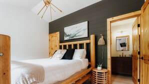 Une chambre à coucher avec un lit en bois, une petite table ronde ornée d’une plante, un téléviseur fixé au mur et un mur de couleur sombre.