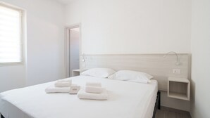 Een minimalistisch hotelkamer met een groot bed, witte lakens, een nachtkastje en een raam met jaloezieën.