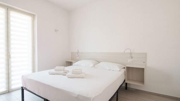 A minimalist bedroom with a bed, two bedside tables, and a large window with blinds. - Das alte Haus - Room 302 (Olbia)