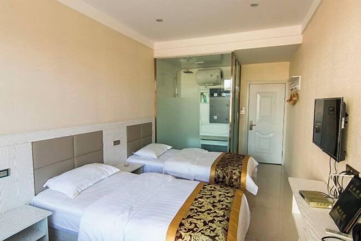 Habitación de hotel con dos camas, televisor, escritorio y baño visible a través de una puerta de cristal.