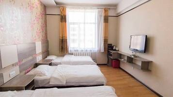 A hotel room with three single beds, a television on a shelf, and a window with curtains.