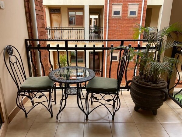 Uma varanda com uma mesa redonda e duas cadeiras, um vaso de planta e vista para um edifício com varandas.