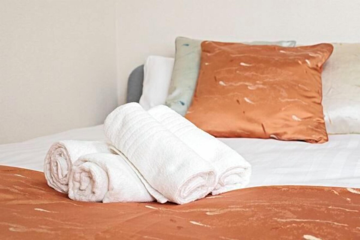 A neatly made bed with rolled white towels and a brown blanket, accompanied by pillows in various colors.