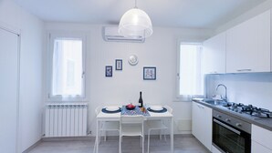 A compact kitchen with white cabinets, a gas stove, a built-in oven, a dining table with chairs, and a pendant light. - Maison De La Brodeuse by Ca-Sa Check (Chioggia)