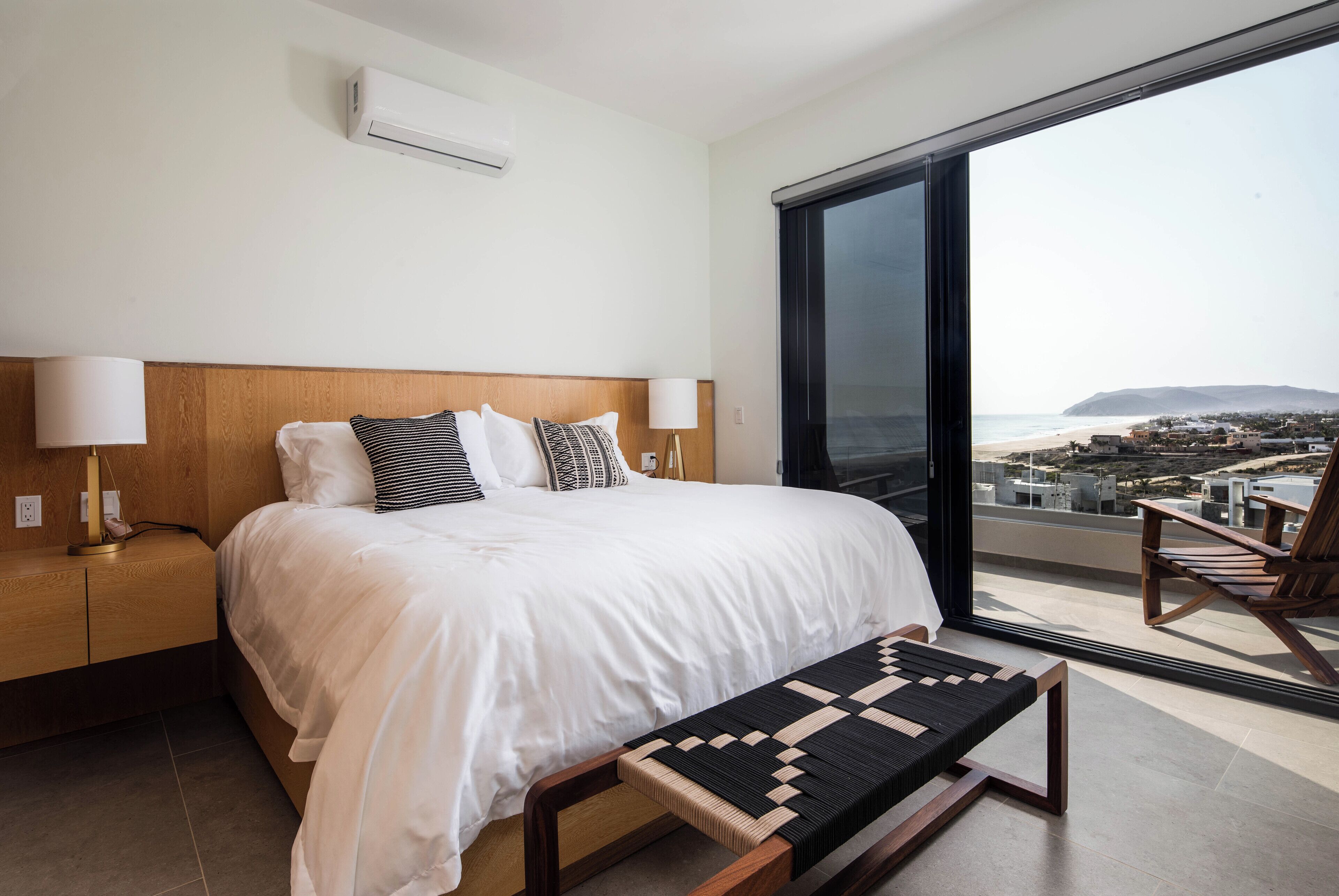 A modern hotel room with a large bed, a bedside table with a lamp, a bench, and a view of the beach through a large window.