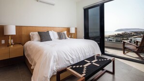Une chambre d’hôtel moderne avec un grand lit, une table de chevet avec une lampe, un banc et une vue sur la plage par une grande fenêtre.