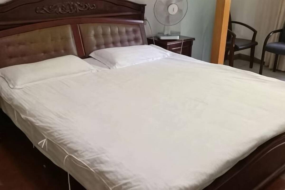 A neatly made bed with a wooden headboard, a bedside table with a lamp, and a chair in the background.