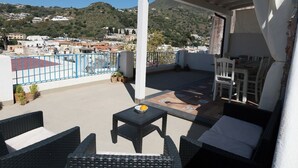 A rooftop terrace with outdoor furniture, a view of a hillside town, and a blue metal railing.