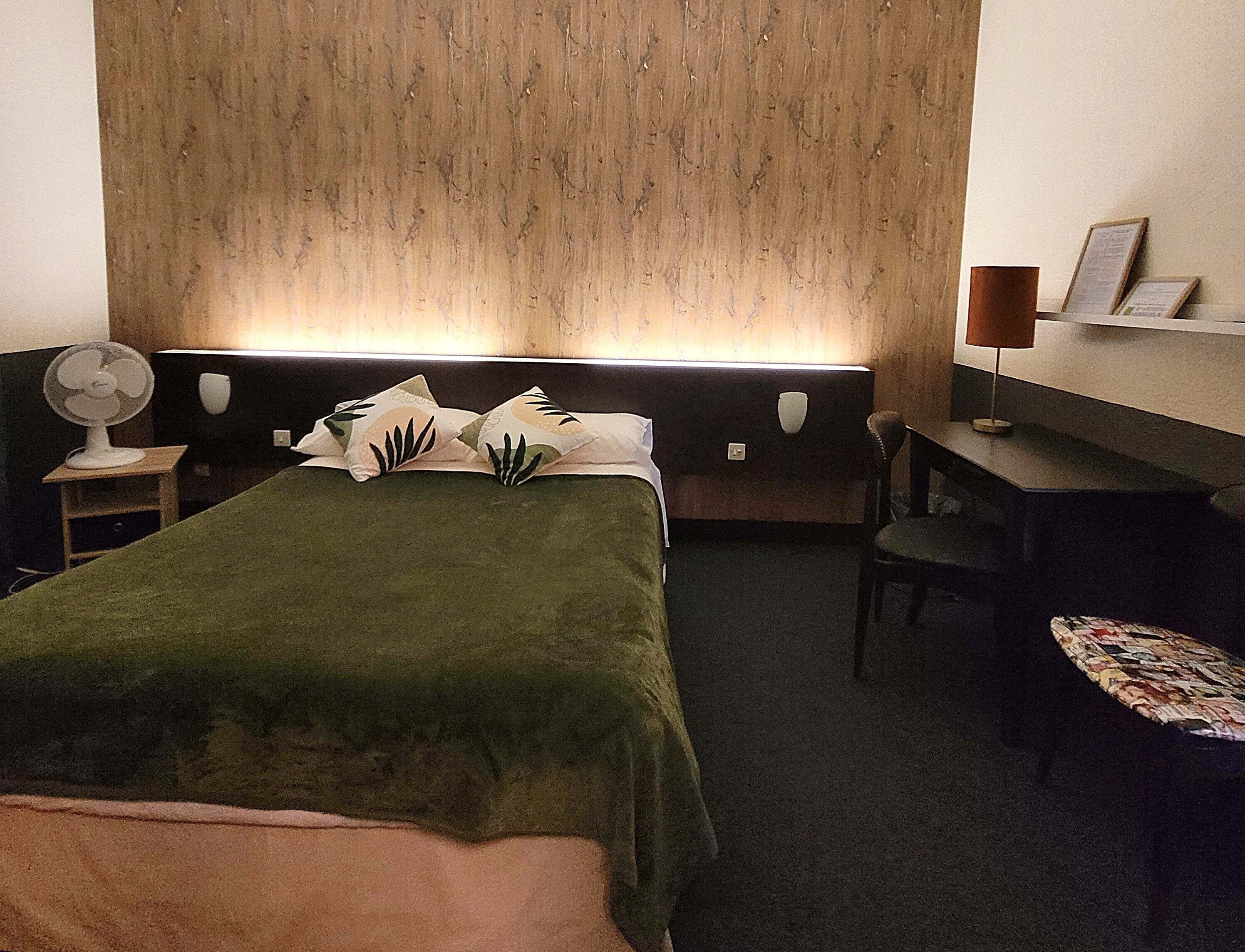 Una habitación de hotel con una cama, un escritorio, una silla, un ventilador y una estantería en la pared.