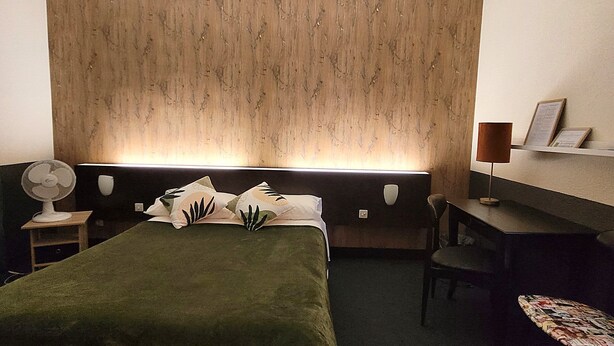 A hotel room with a bed, a desk, a chair, a fan, and a wall-mounted shelf. - LOGIS Hôtel Le Merle Blanc (Digoin)
