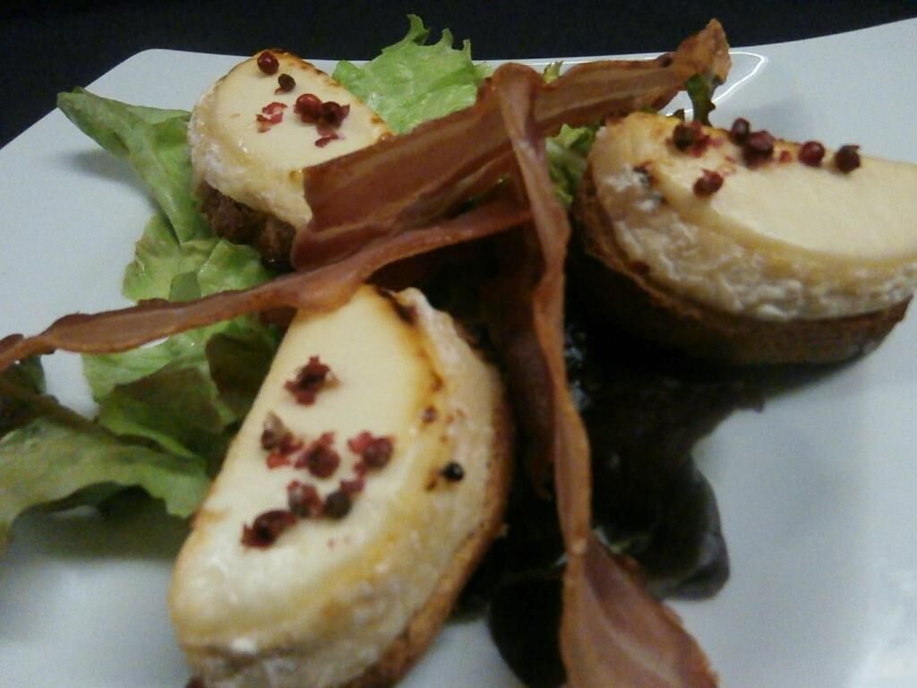 A plate with a dessert consisting of a creamy topping, possibly cheesecake, garnished with red bits, accompanied by a dark sauce and green leafy vegetables.
