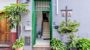 A building entrance with a green door, a wooden door, and a staircase leading inside. There are potted plants outside and a cross on the wall.