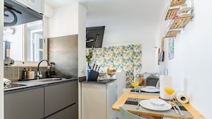 A compact kitchen with a sink, stove, and a small refrigerator. There's a dining area with a table set for two, a chair, and a vase with flowers.