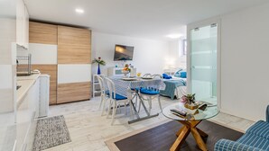 Una sala de estar moderna y bien iluminada, con comedor, televisor y puerta de vidrio que da al dormitorio.