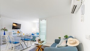 Una sala de estar moderna con un sofá azul, un comedor con mesa y sillas, un televisor montado en la pared y una puerta de cristal que da a otra habitación.