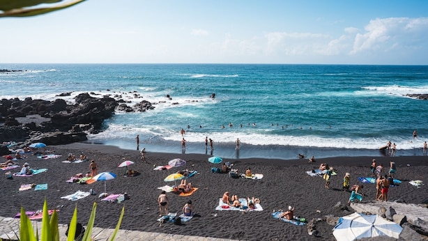 Una playa con gente tomando el sol y nadando, costa rocosa y mar azul cristalino.