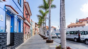 Calle bordeada de palmeras y edificios de colores, una camioneta blanca estacionada al lado, y un cielo despejado.