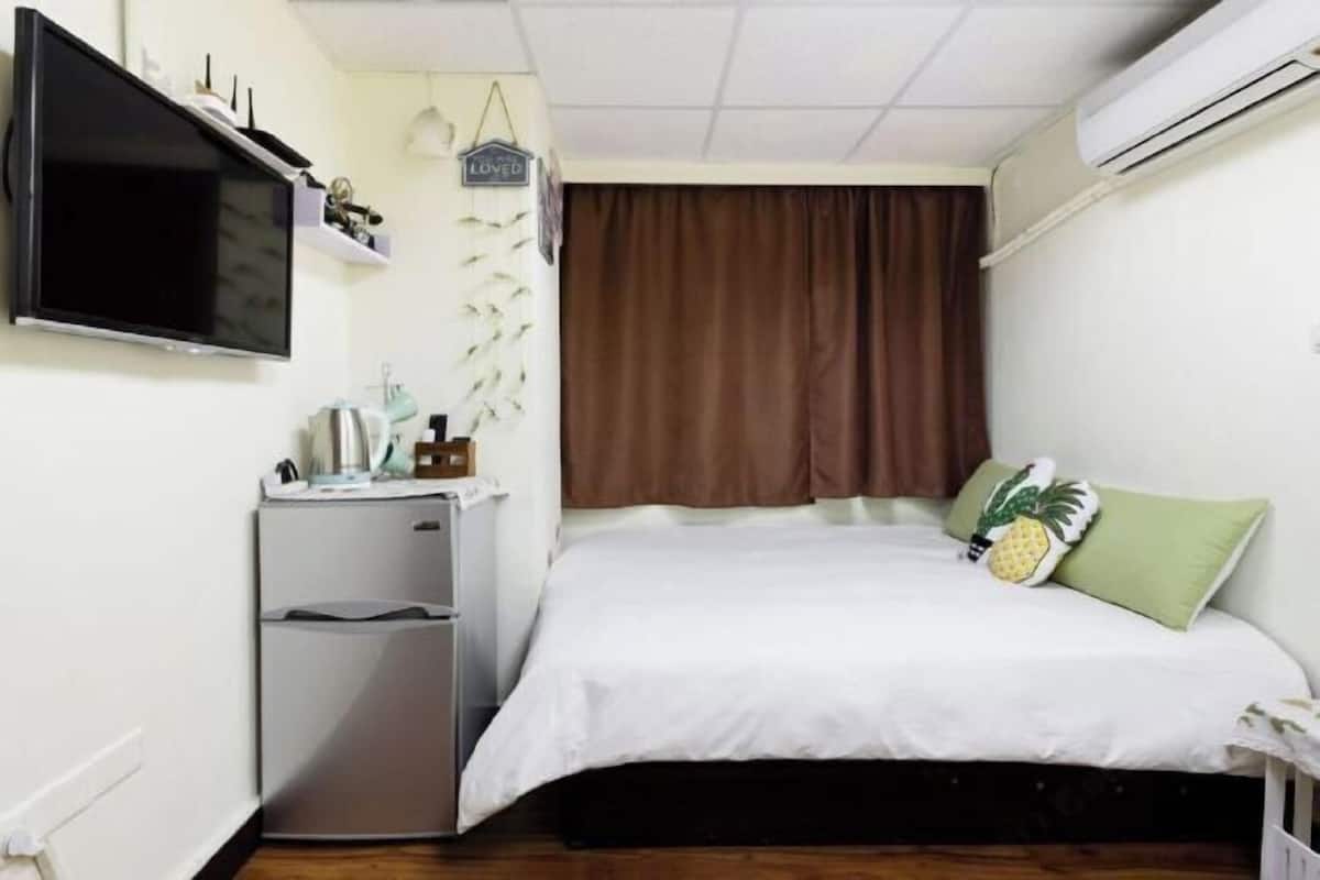 A bedroom with a bed, a TV mounted on the wall, a small refrigerator, and a window with curtains.