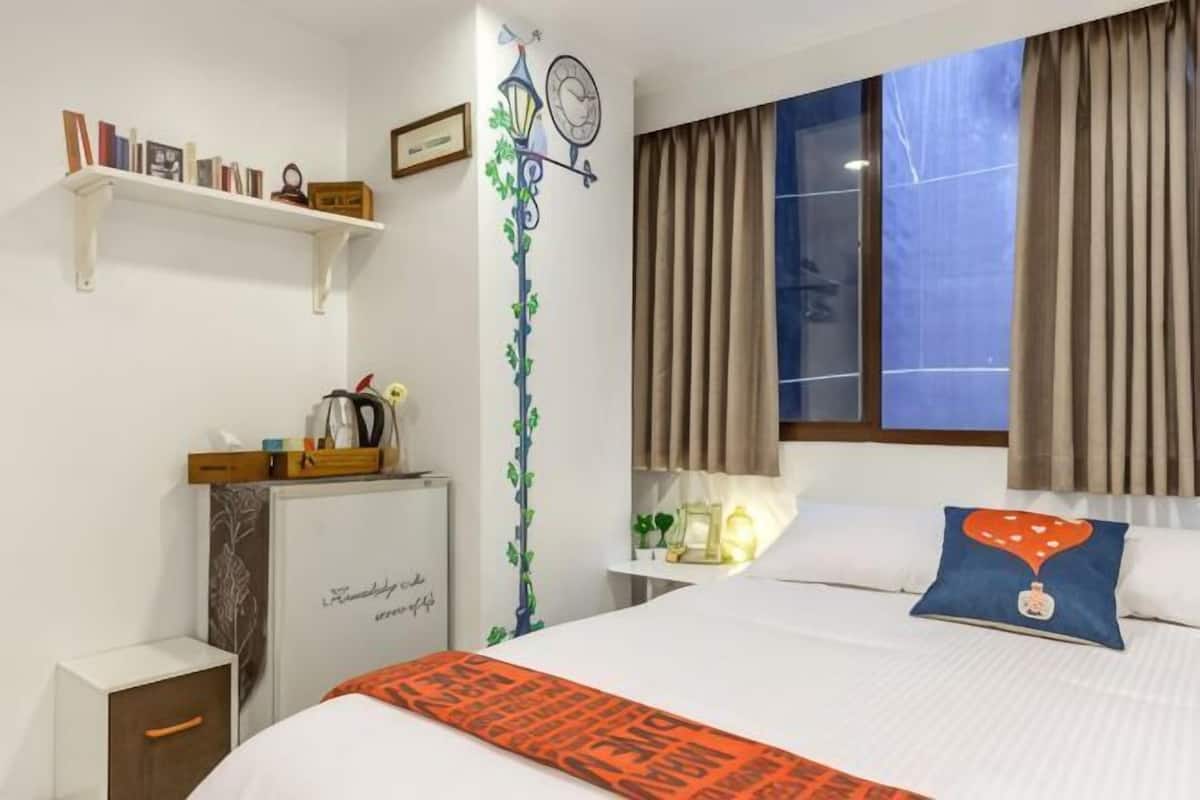 A hotel room with a bed, a desk, a shelf with books, a decorative wall, and a window with curtains.