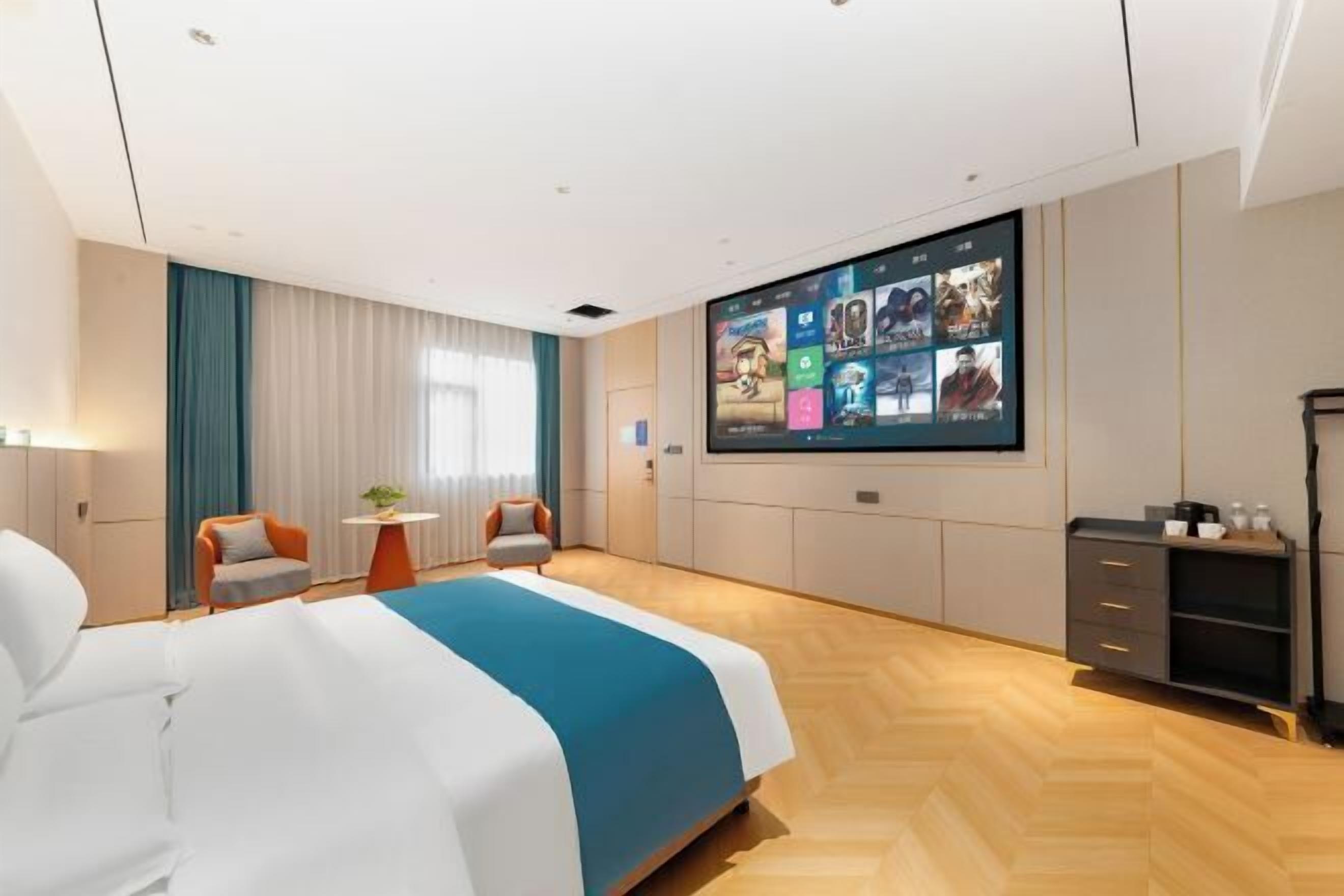 A modern hotel room with a large bed, a seating area with two chairs, a small table, a TV mounted on the wall, and a wardrobe.