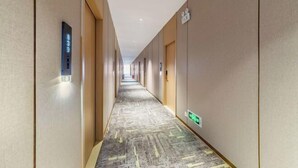 Un lungo e stretto corridoio di un hotel con pavimento rivestito di moquette, un display digitale dell'ascensore e un cartello verde di uscita di sicurezza.