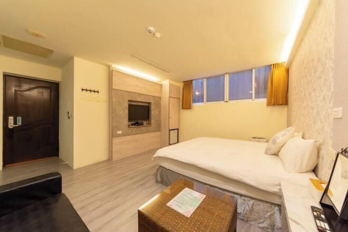 A hotel room with a bed, a small table with a paper, a TV, and a window with curtains.