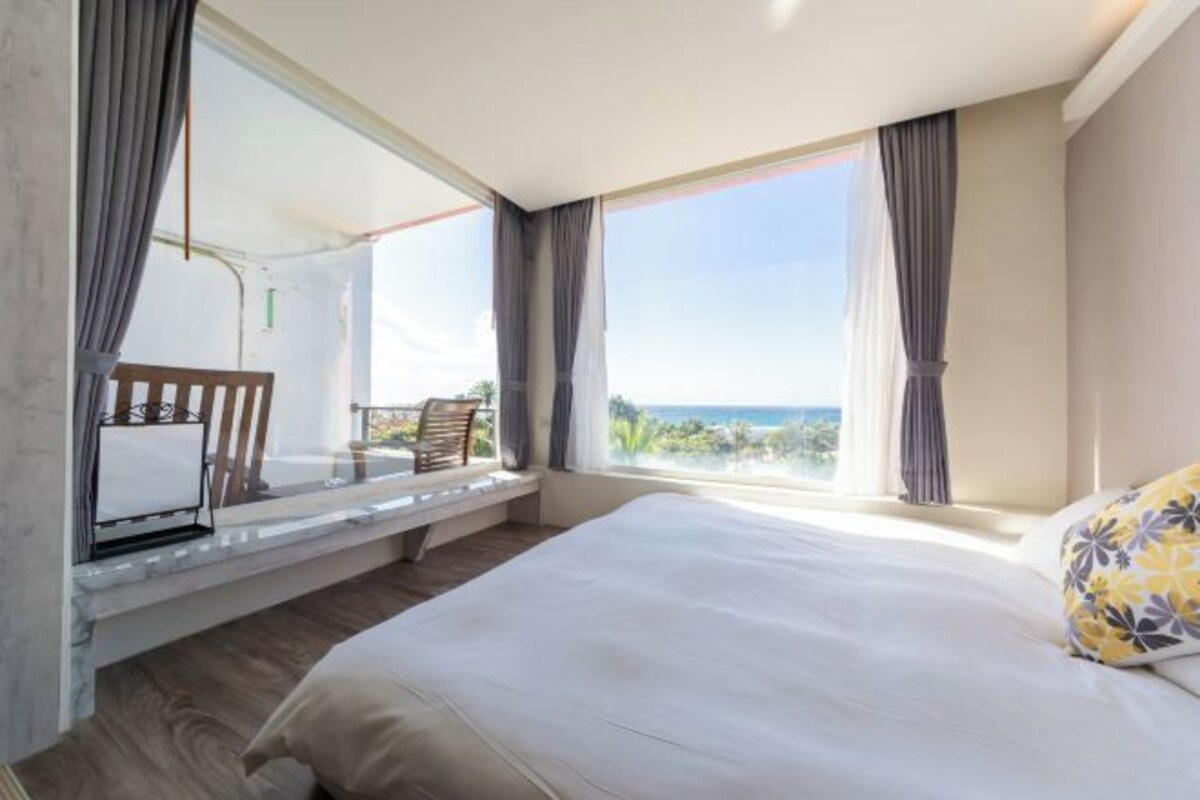 A bedroom with a large bed, a window seat with a chair, and a view of the outdoors.