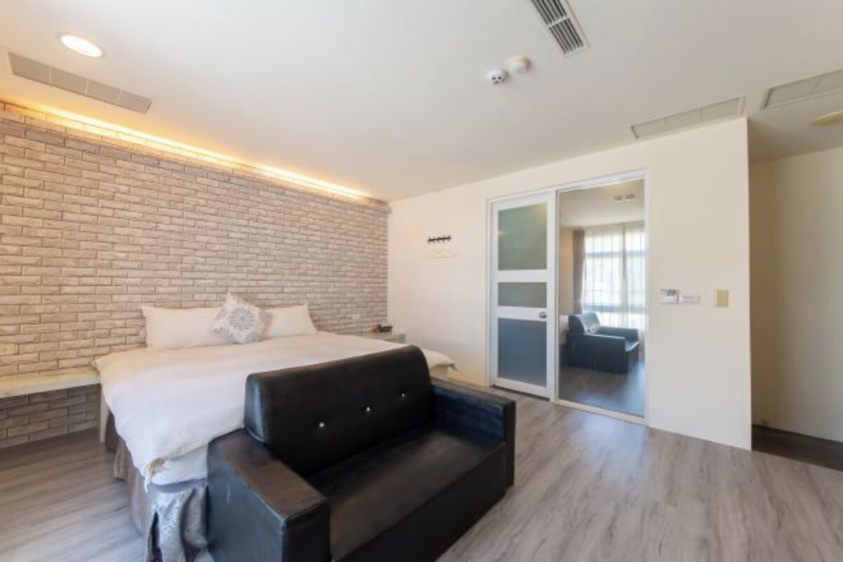 A modern hotel room with a brick accent wall, a large bed, a black sofa, and a sliding glass door leading to a balcony.